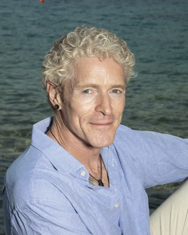 Phil Duval by the water in a blue linen shirt, smiling at the camera